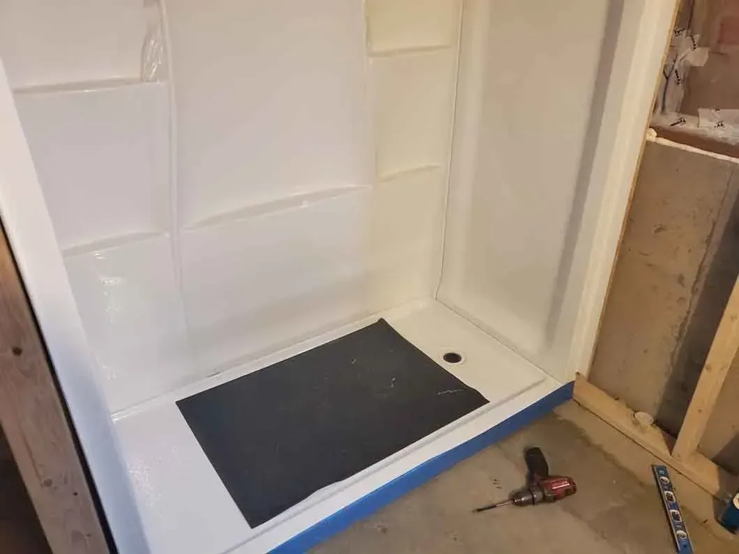 White shower stall with a black floor mat; tools and wood framing suggest a bathroom remodel