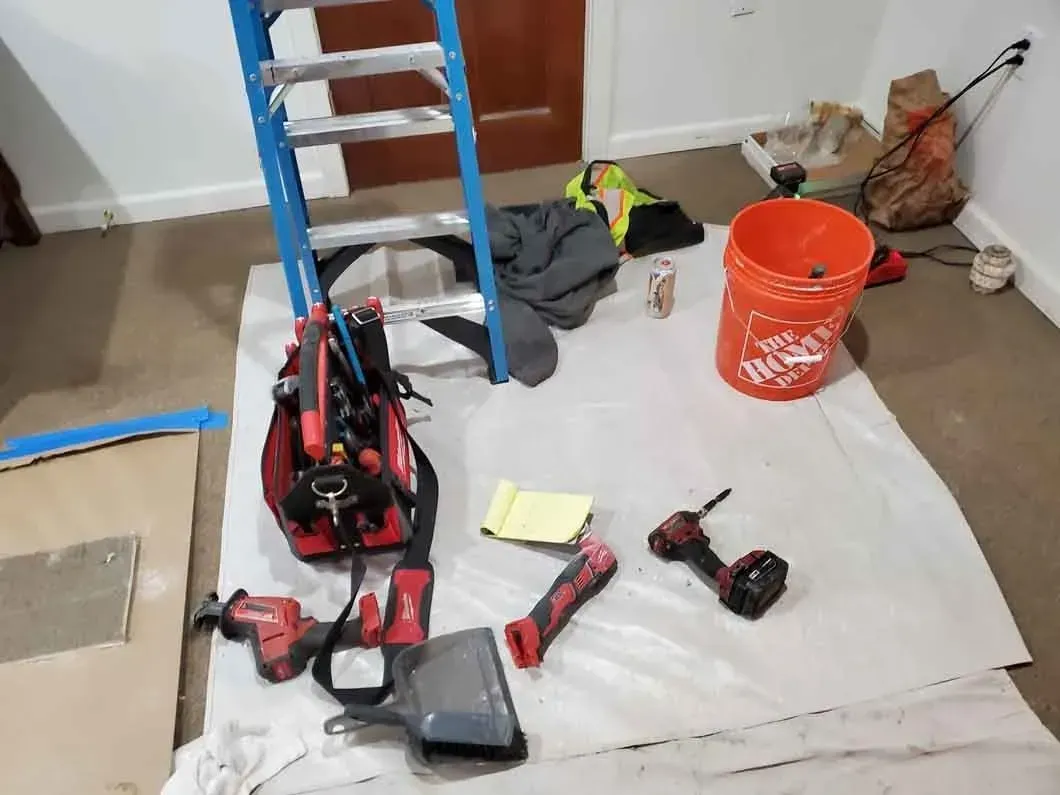 Work area with ladder, tools, orange bucket, and white tarp on a carpeted floor