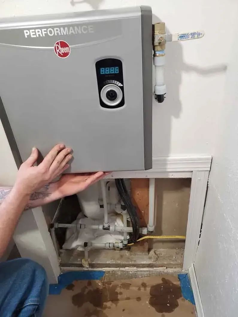 Hand reaching under an open gray utility panel revealing exposed pipes and a wet floor stain.