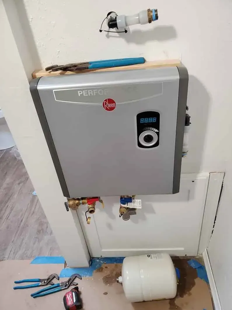 Wall-mounted gray utility box with pipes and fittings, above a white container on the floor in a utility room
