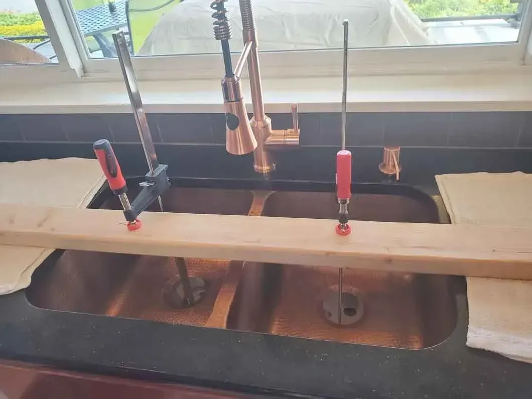 Kitchen sink with faucet and two red clamps holding a wooden board across it under a window