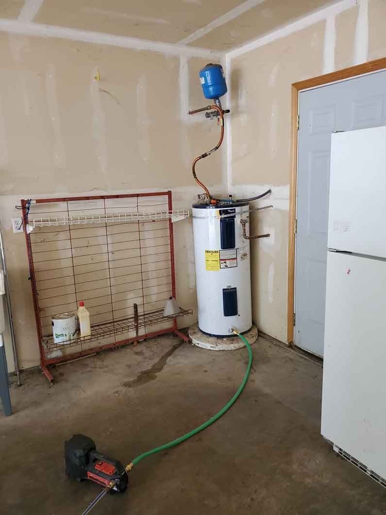 Utility room with water heater, green hose, wall-mounted tank, and white appliance beside a garage door.