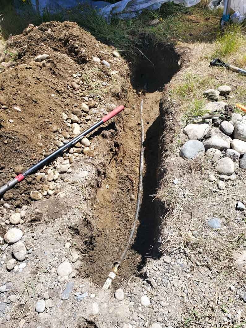 Narrow trench dug in rocky dirt beside a red-and-black pipe under bright sunlight