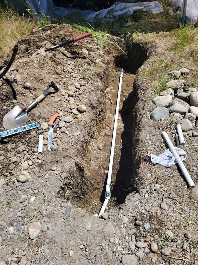 Trench dug in rocky ground with exposed white pipes and scattered tools at the sides.
