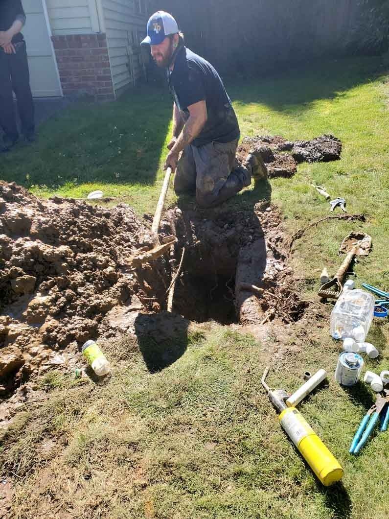 Man digging a large hole in a yard with a shovel, dirt piled nearby and tools scattered around