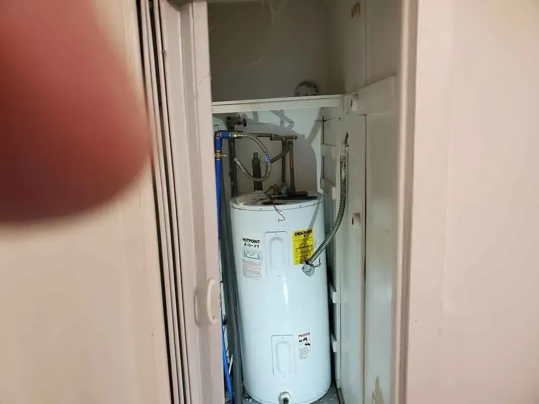 White water heater in a narrow utility closet with pipes and a finger partially blocking the left side