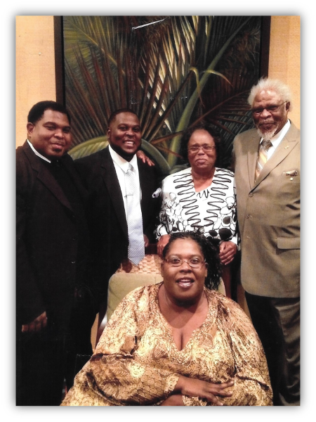 Five people pose for a group photo in front of a large plant, wearing formal attire and smiling at the camera.