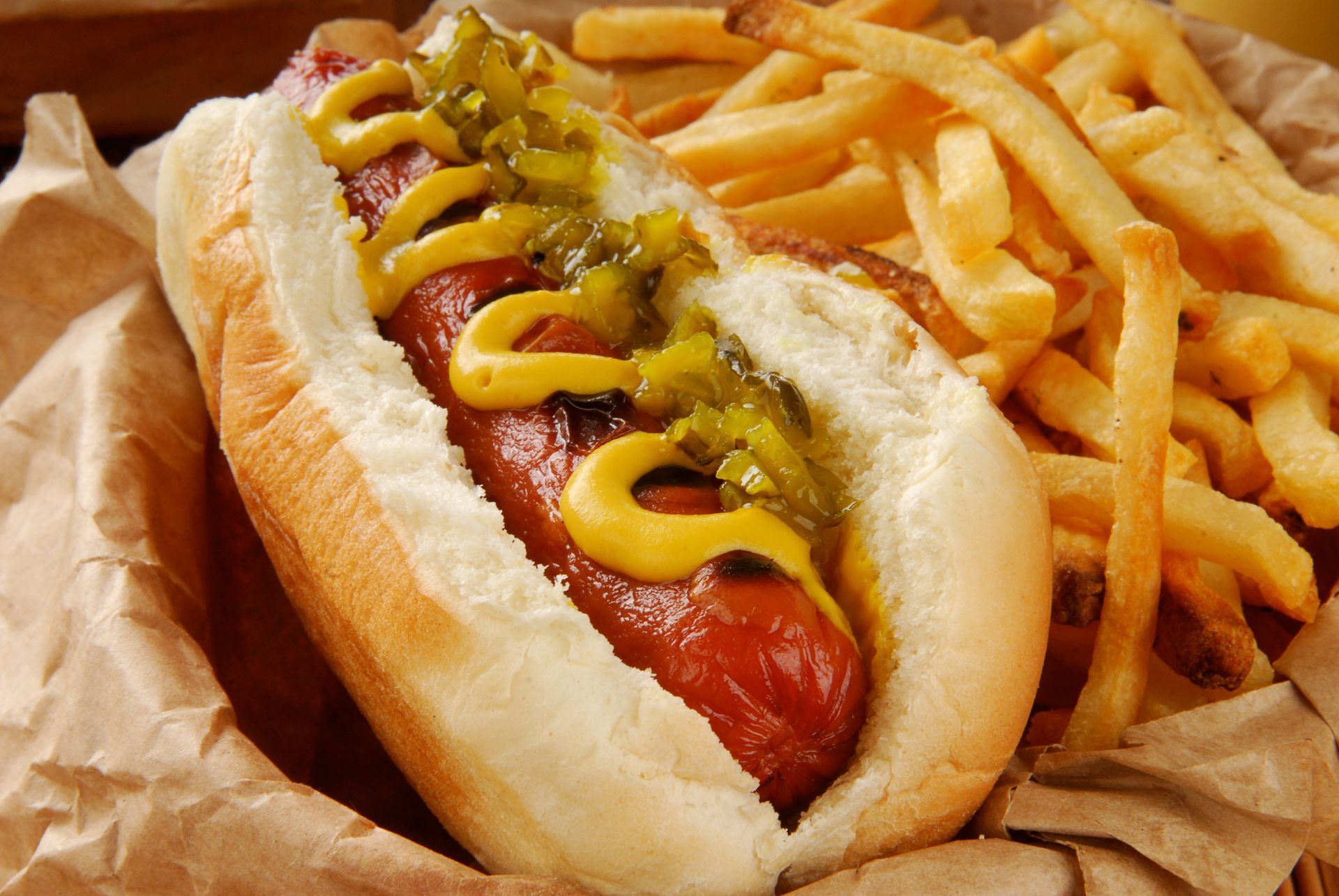 Hot dog with mustard and relish beside a pile of French fries on brown paper.