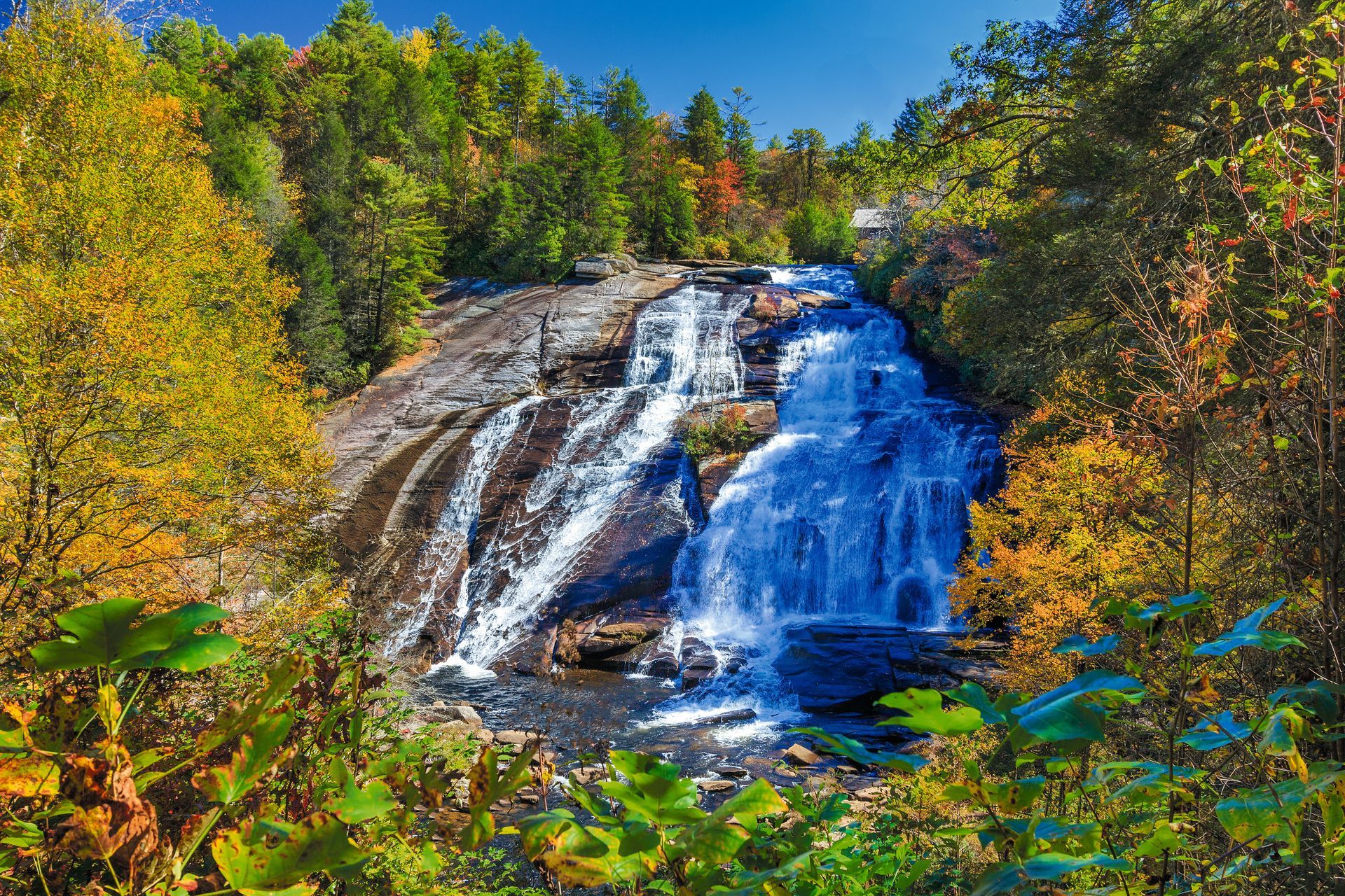 Waterfall cascading over dark rocks in a forest, surrounded by bright green and autumn-colored trees.