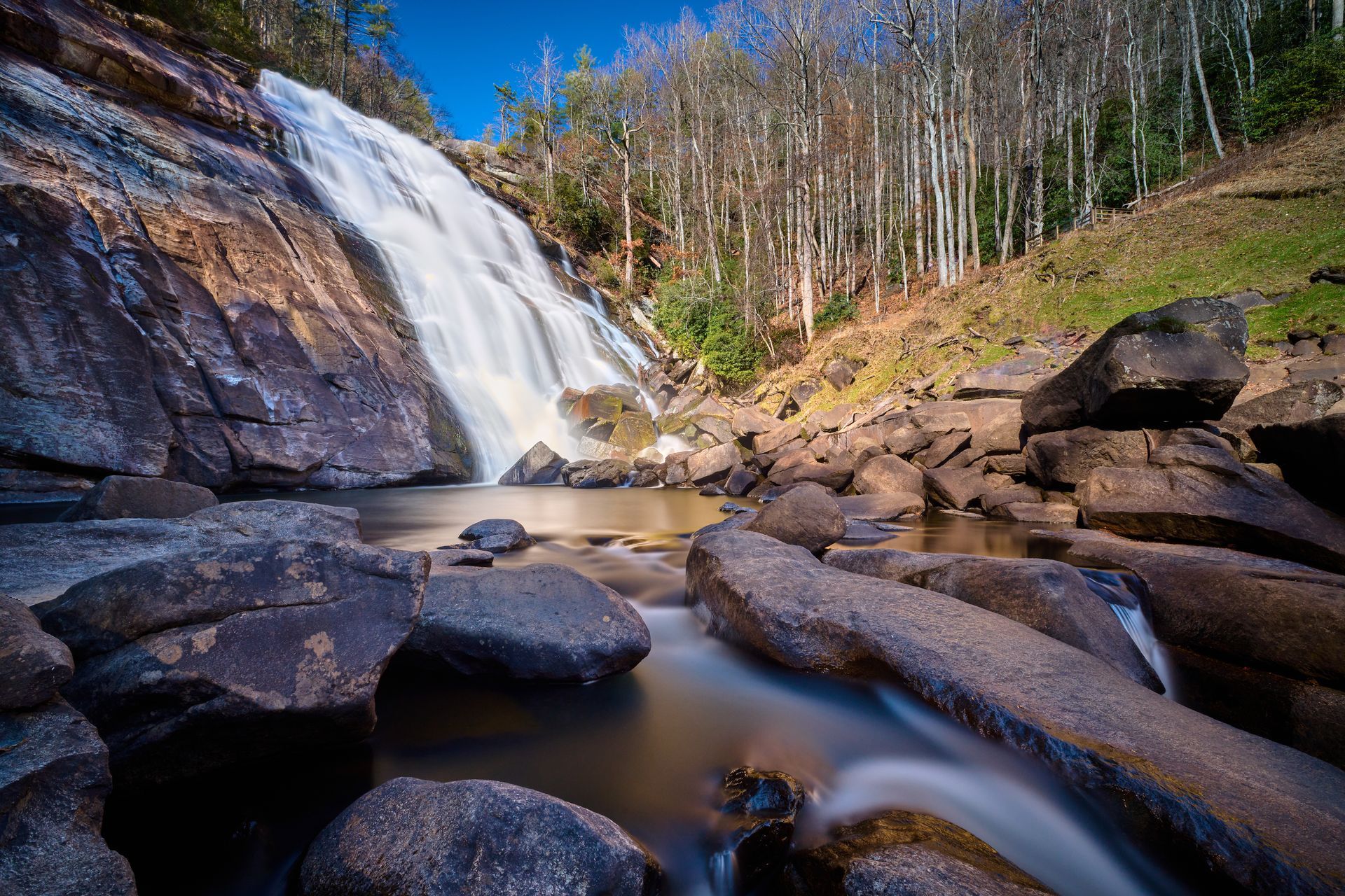 Waterfall cascading over rocks into a calm pool, surrounded by forest and blue sky.