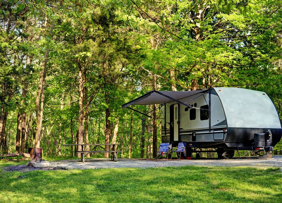 Small camper trailer with awning in a green forest campsite under tall trees