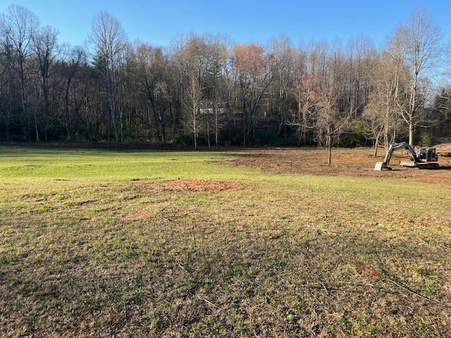 Grassy field with trees under a clear blue sky; a small excavator sits at the right edge.