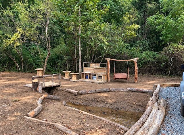 Outdoor campsite with wooden benches, a fire pit, and a simple shelter in a forest clearing