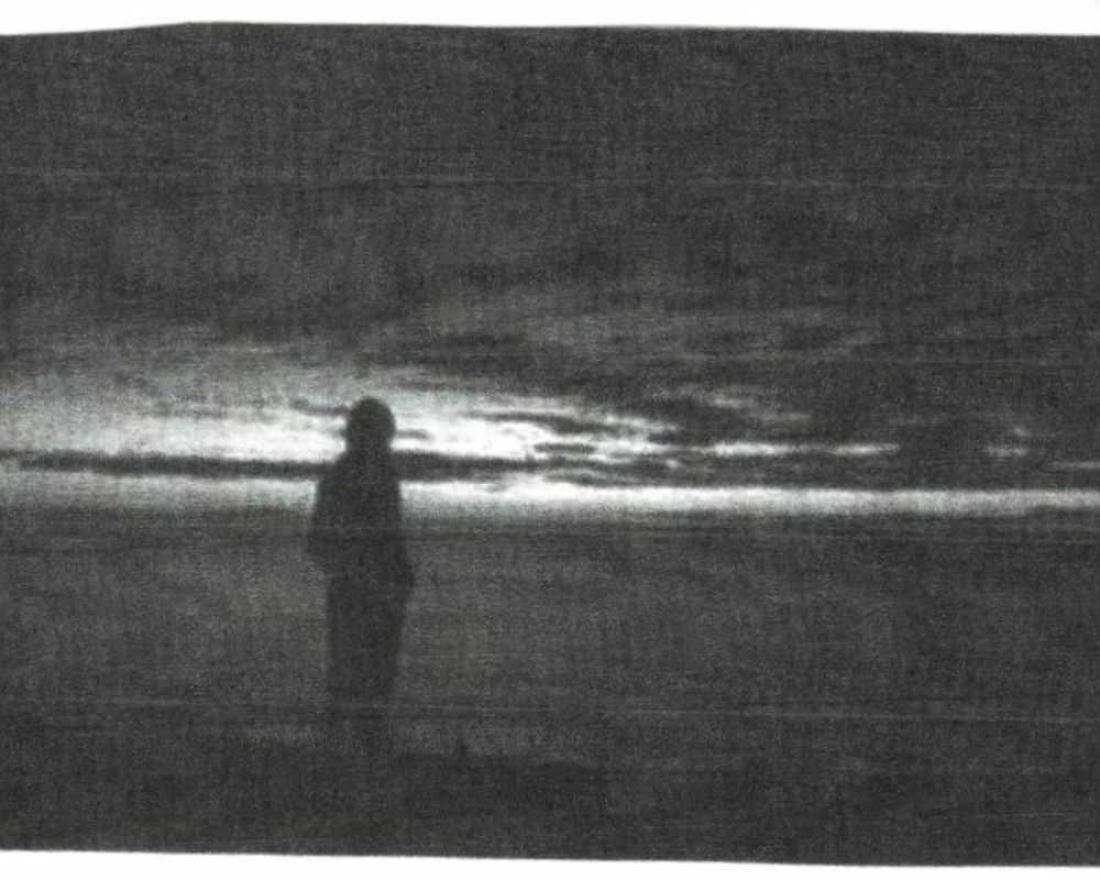 A black and white photo of a person standing on a beach at sunset.