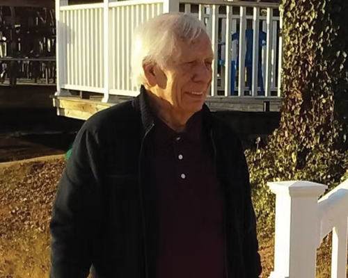 Rob wearing a black jacket is standing in front of a white fence.