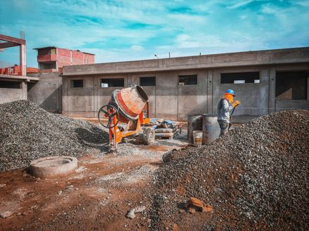 A construction worker in safety gear stands near a concrete mixer and piles of gravel at an active building site.