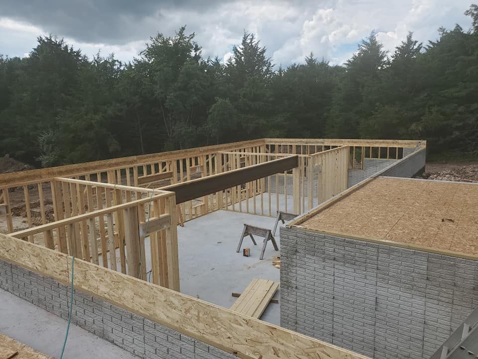 A construction site showing wood framing atop a concrete foundation for a house under development near a tree line.