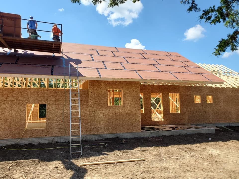 Construction workers on a scaffold install sheathing on the roof of a house under a clear blue sky.
