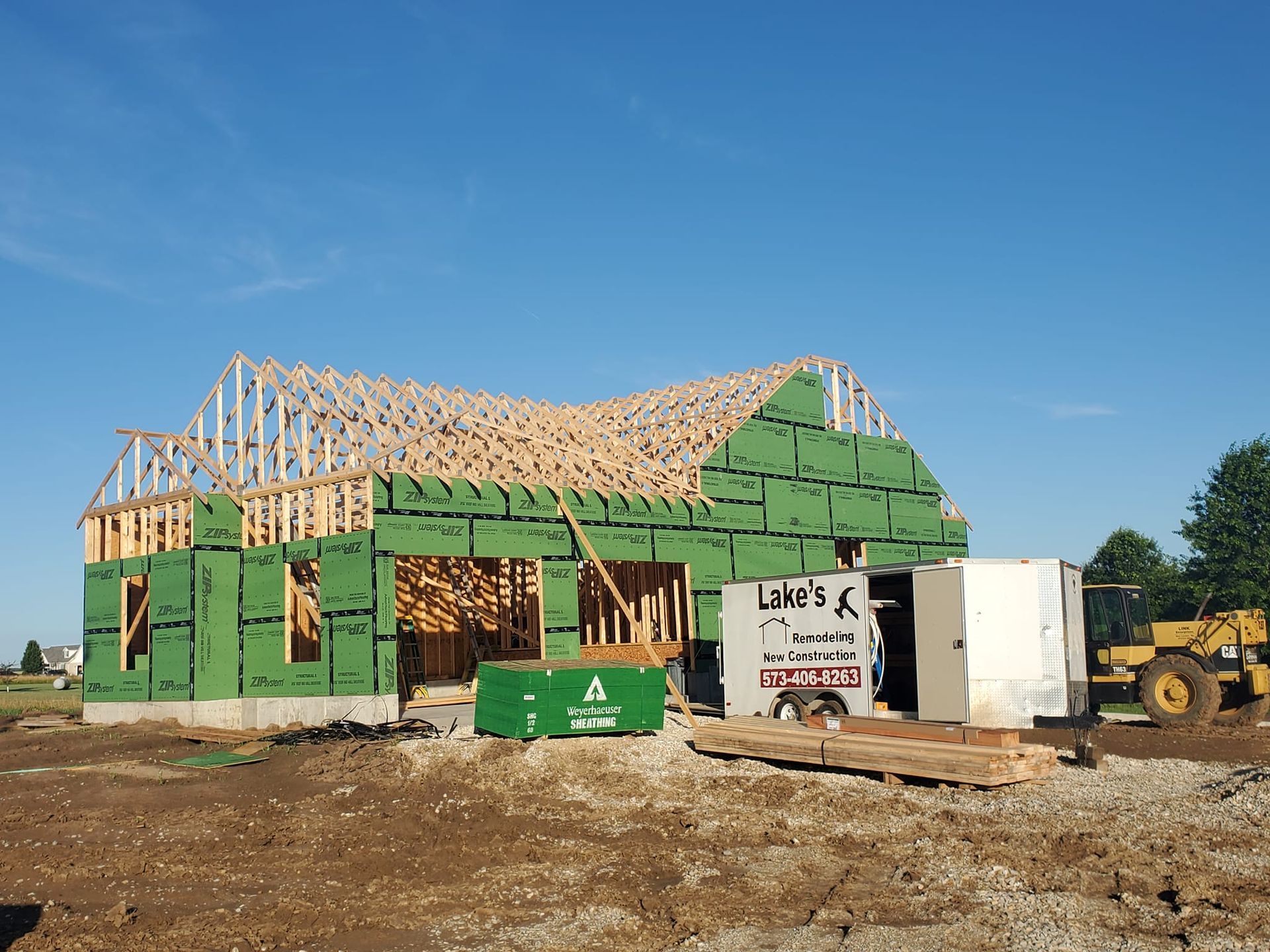 A wooden residential house frame under construction, featuring green sheathing panels under a clear blue sky.
