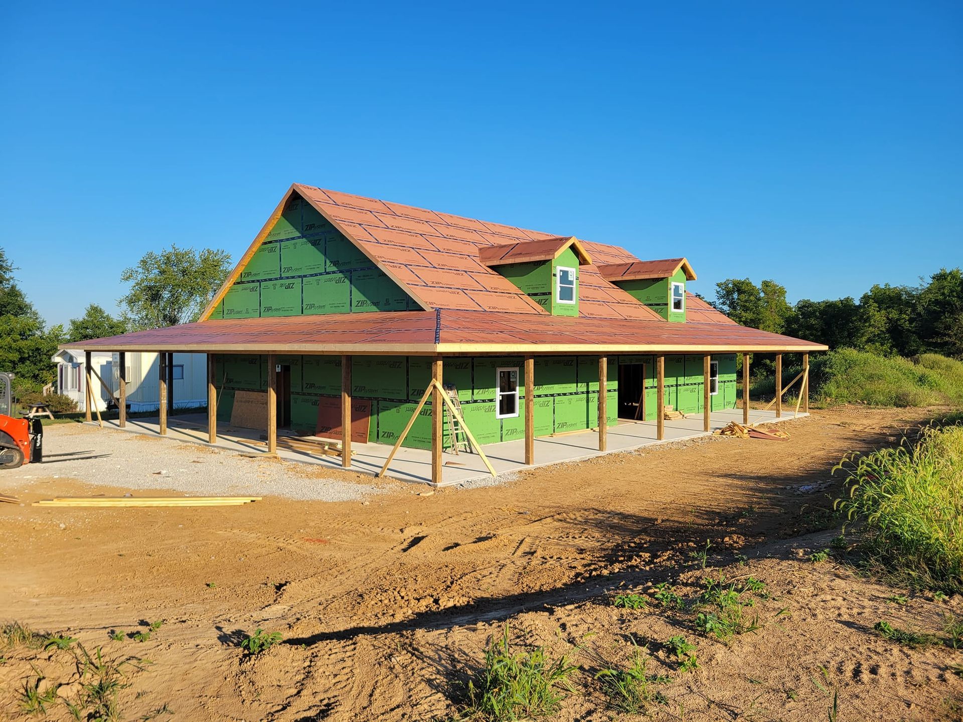 A new house under construction with green exterior sheathing, a red shingled roof, and a wrap-around porch in a rural yard.