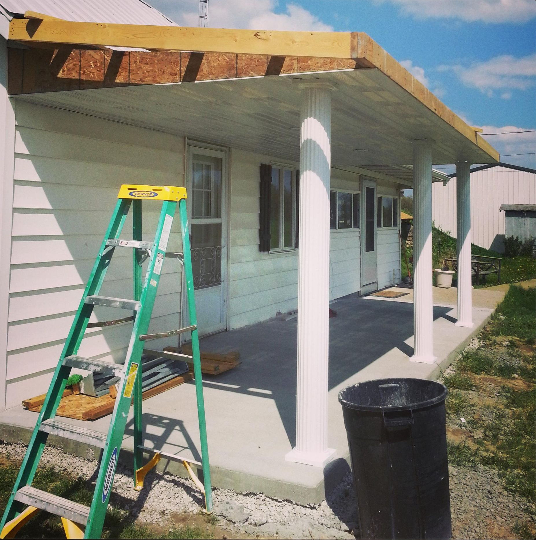 A light-colored house with a newly constructed patio roof supported by white columns, with a green ladder in the foreground.