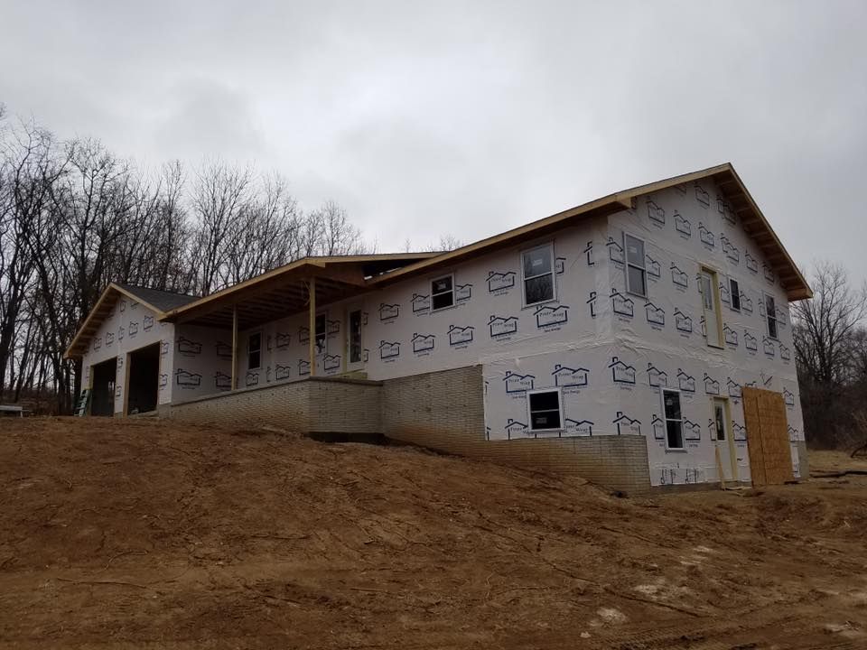 A two-story house under construction with white exterior house wrap, a concrete foundation, and an unfinished porch.