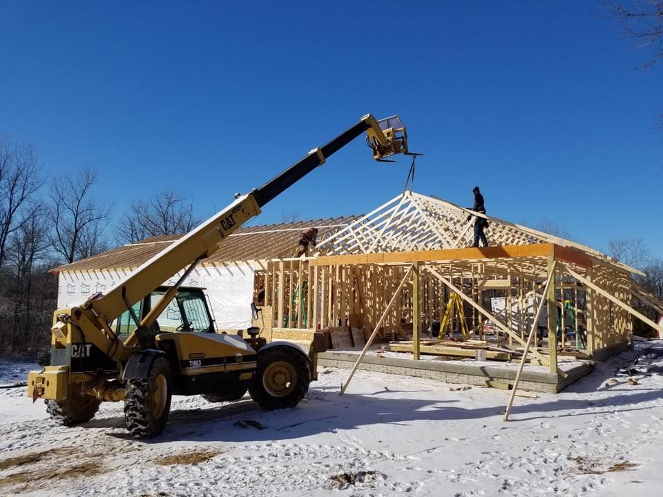 A yellow telehandler lifts a roof truss to workers framing a wood structure on a snow-covered building site.