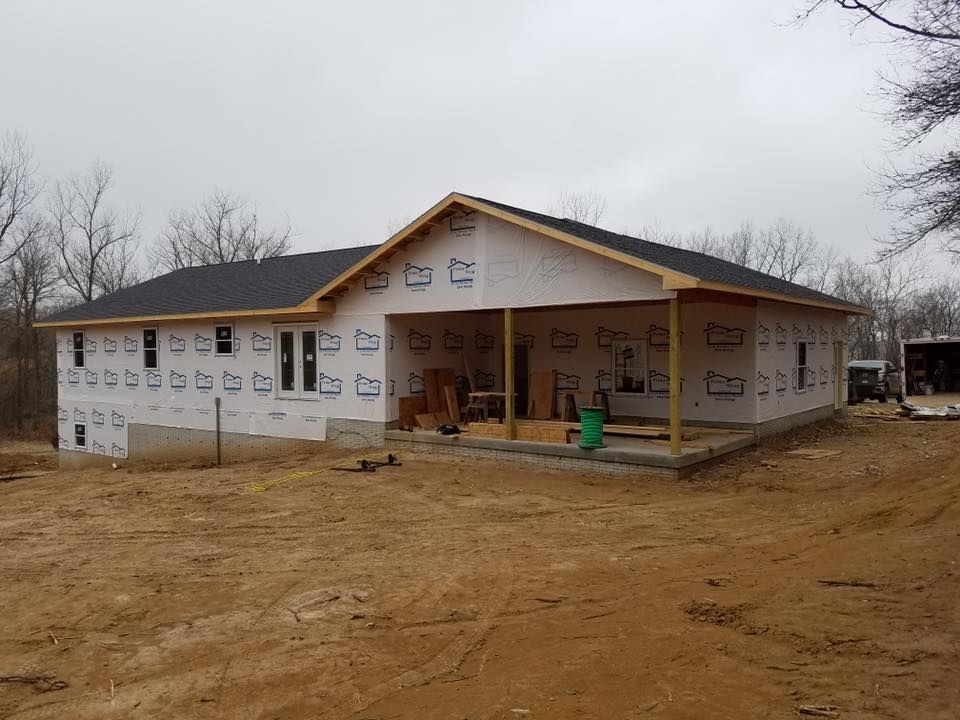 A house under construction with white protective wrap on the walls, a dark shingled roof, and an open front porch.