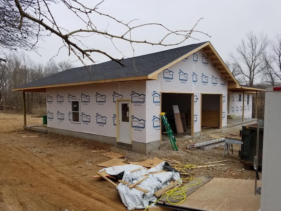 An unfinished garage with a dark shingled roof, white house wrap exterior, and an open entryway on a dirt lot.