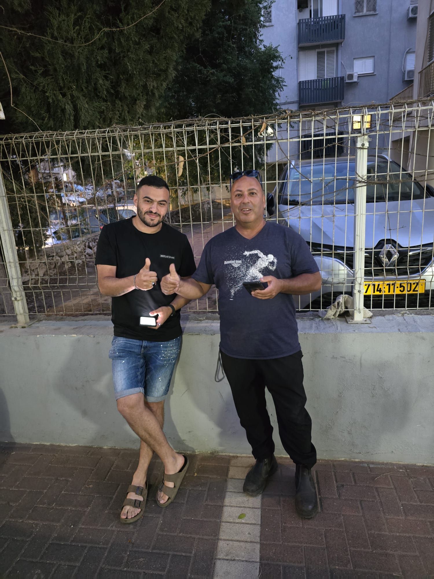 שני אנשים מצטלמים עם משקאות בלילה ליד גדר זהב ובניין דירות.