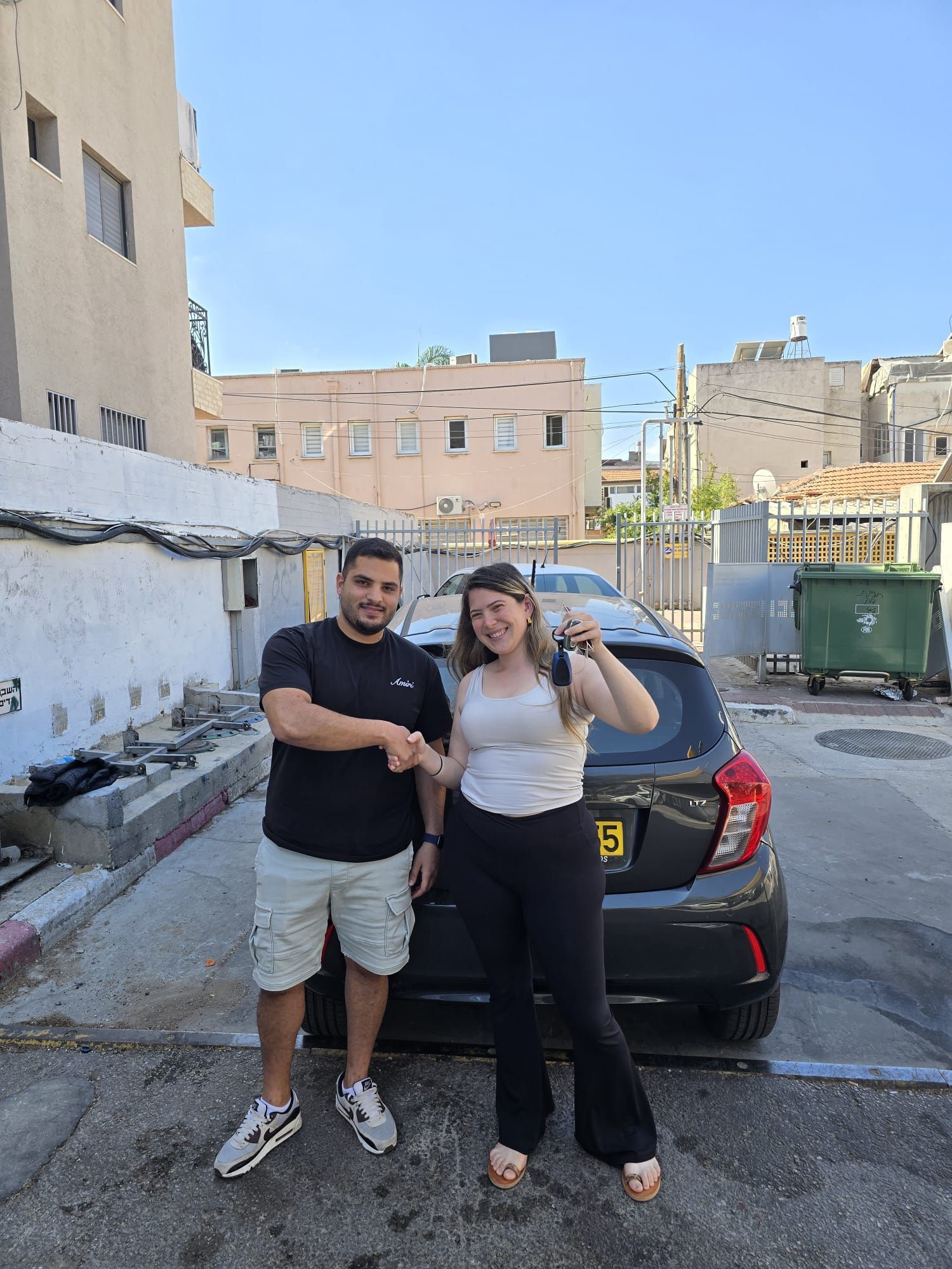 שני אנשים מצטלמים ליד מכונית שחורה בחניון, כשמאחוריהם בניינים ופח אשפה ירוק.