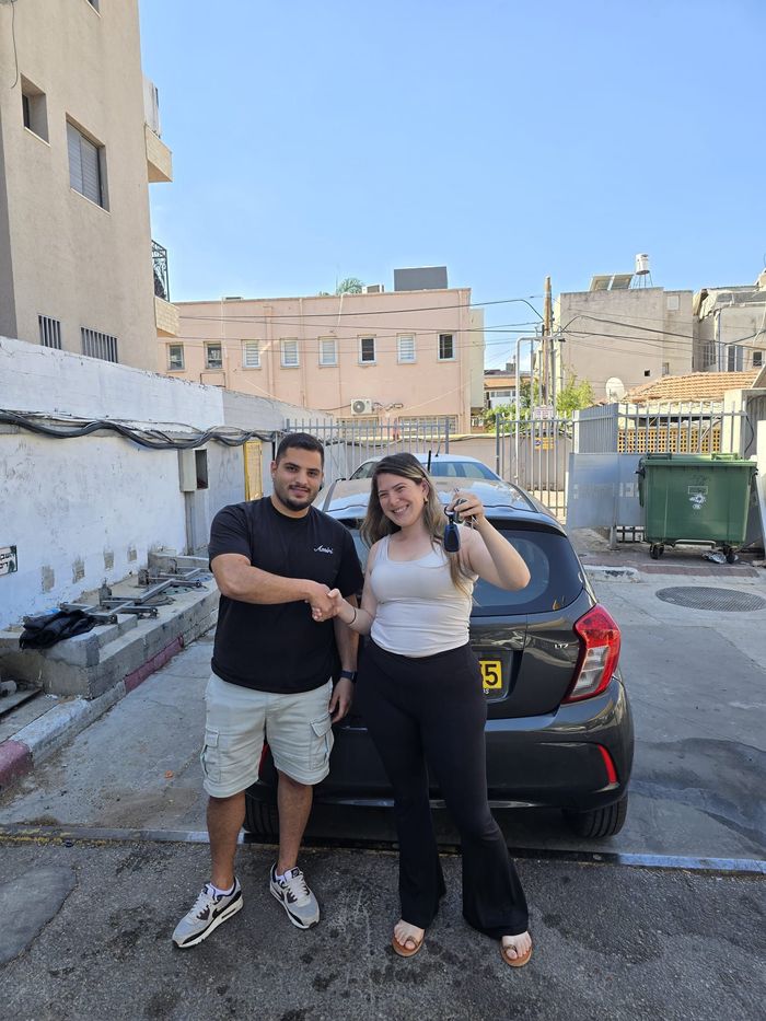 שני אנשים מצטלמים ליד מכונית שחורה בחניון, כשמאחוריהם בניינים ופח אשפה ירוק.
