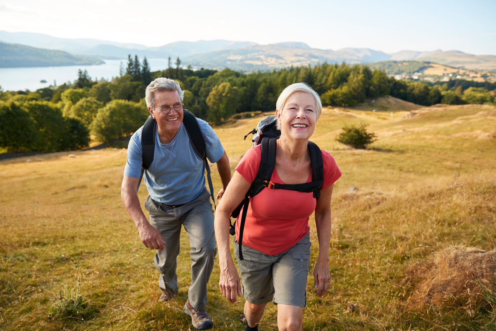 Senior couple smiling while hiking up a grassy hill with a lake and mountains in the background.