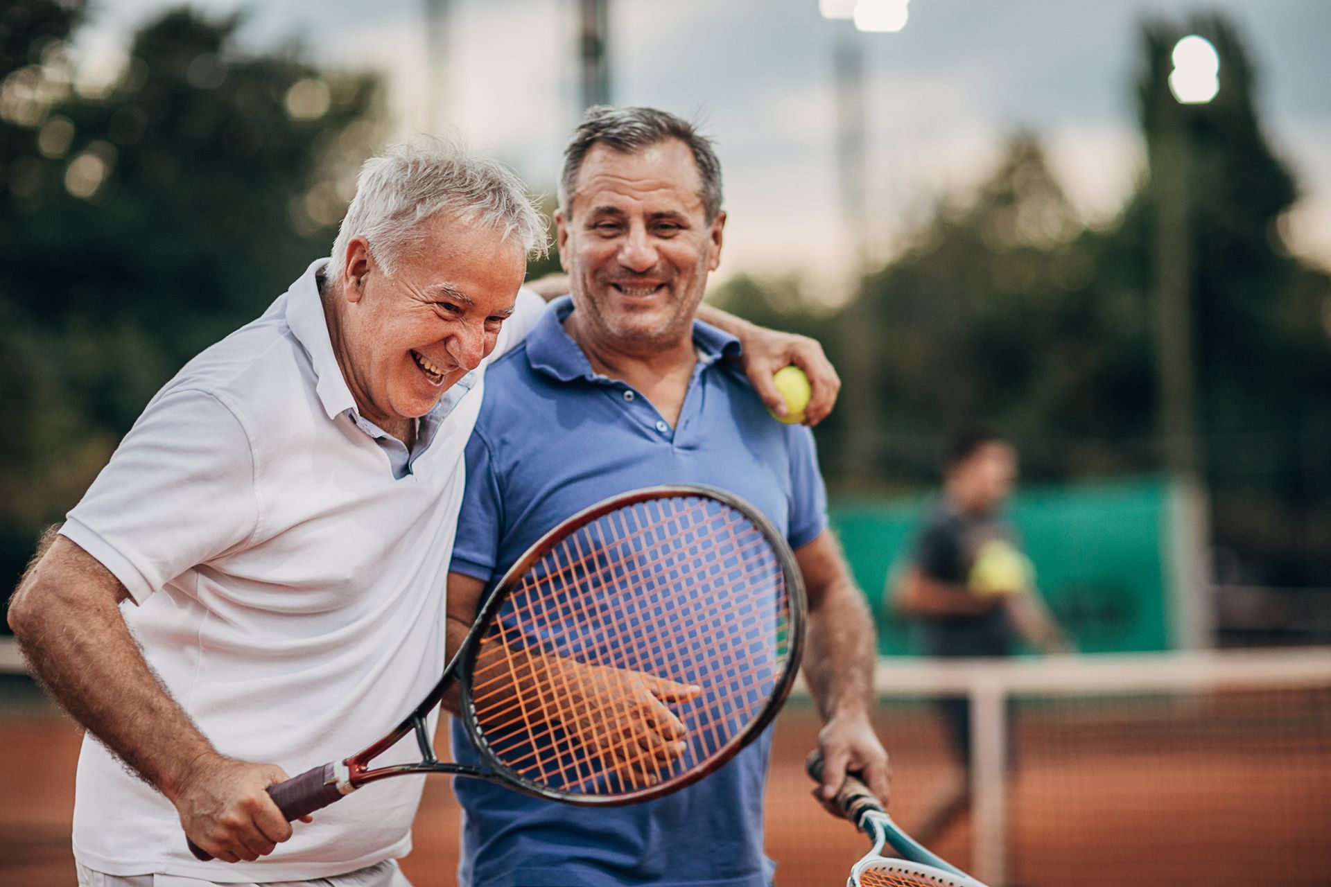 Two men smiling on a tennis court, one with arm around the other holding a racket and ball.