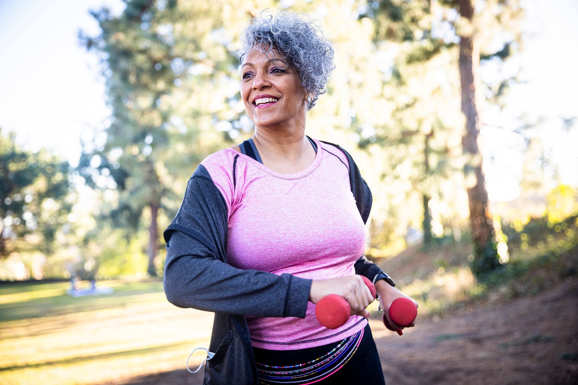 Woman with gray hair smiles while exercising outdoors with red weights.