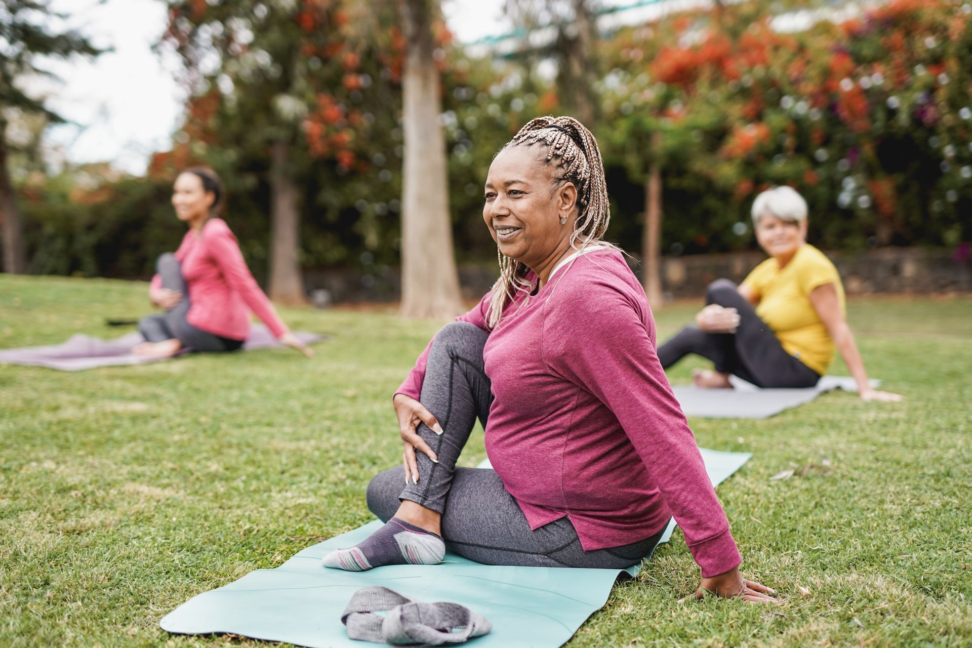 Three women doing yoga outdoors on mats, smiling and stretching in a grassy park.