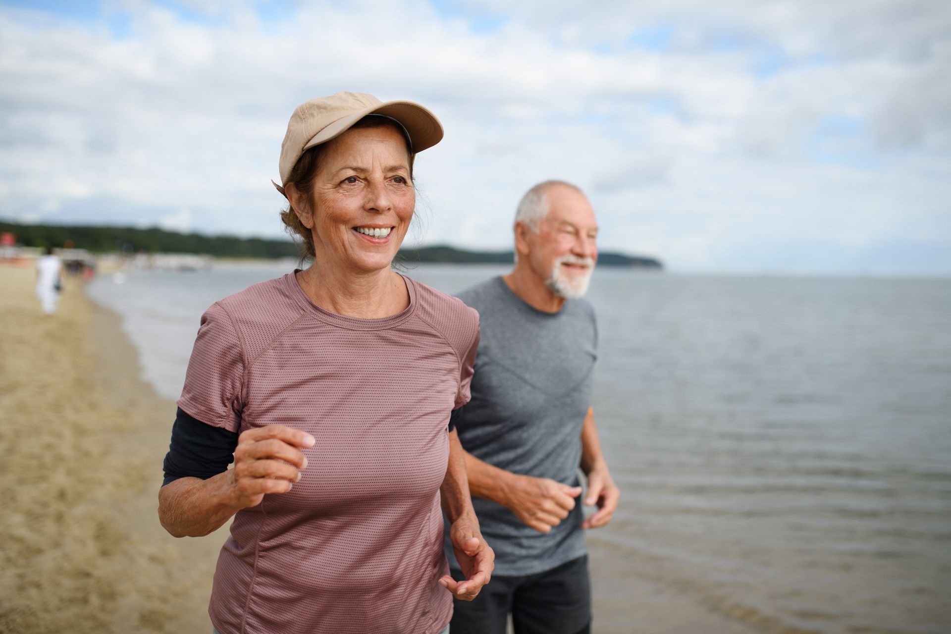 Older couple jogging on a beach, smiling, under cloudy sky.