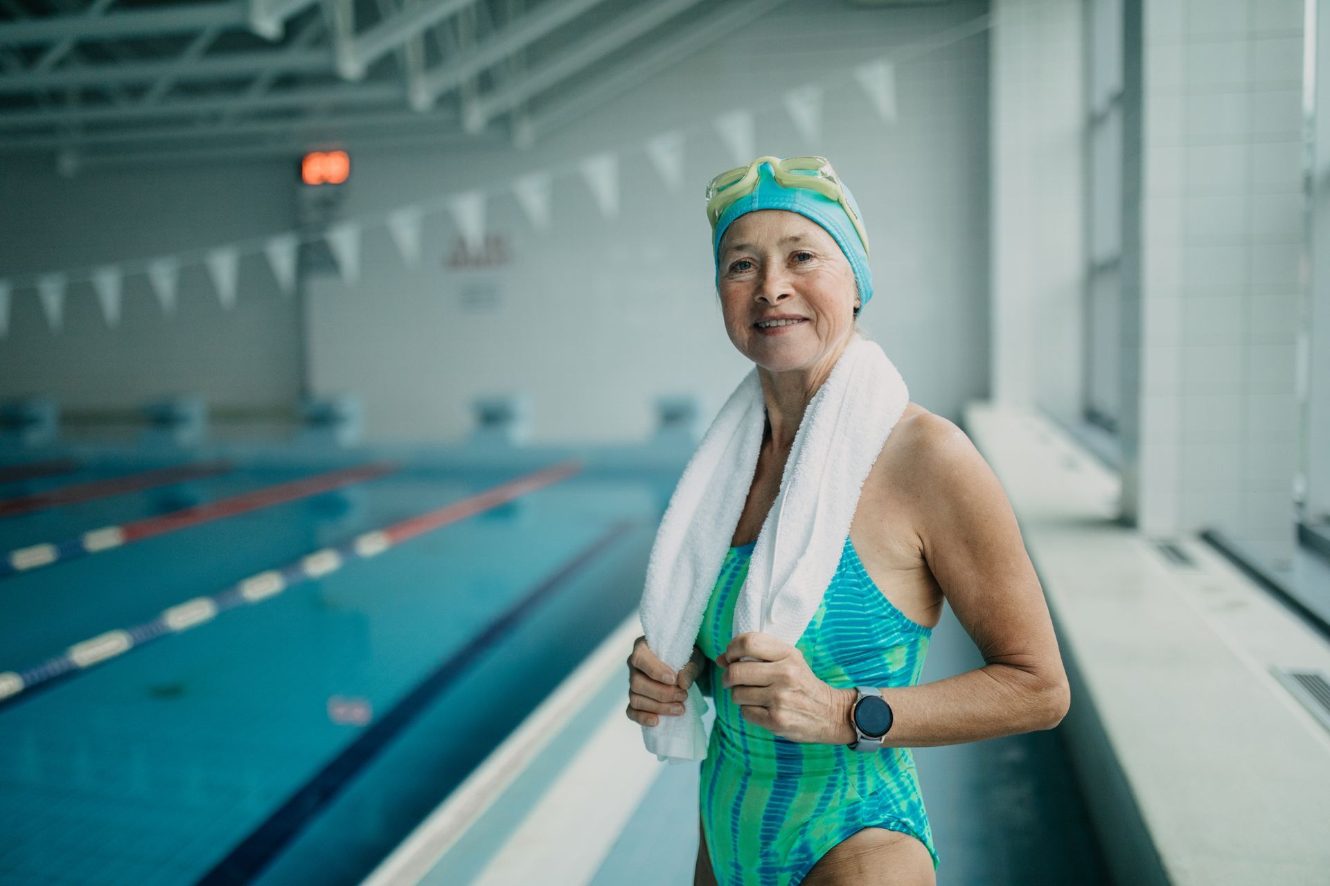 Woman in a swimsuit and swim cap by a pool, holding a towel and smiling.
