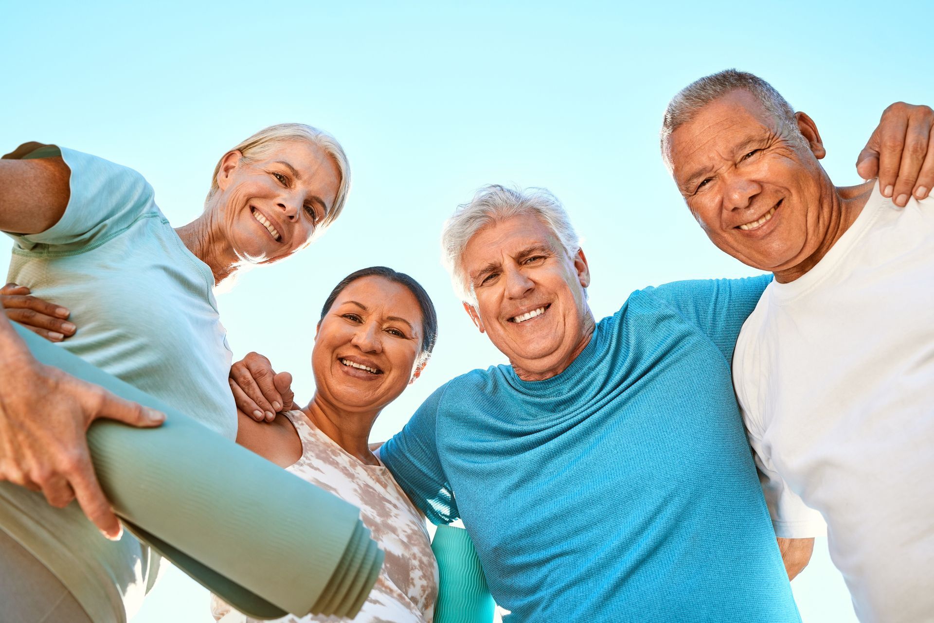 Four diverse seniors smile and embrace, one holds a yoga mat, under a blue sky.