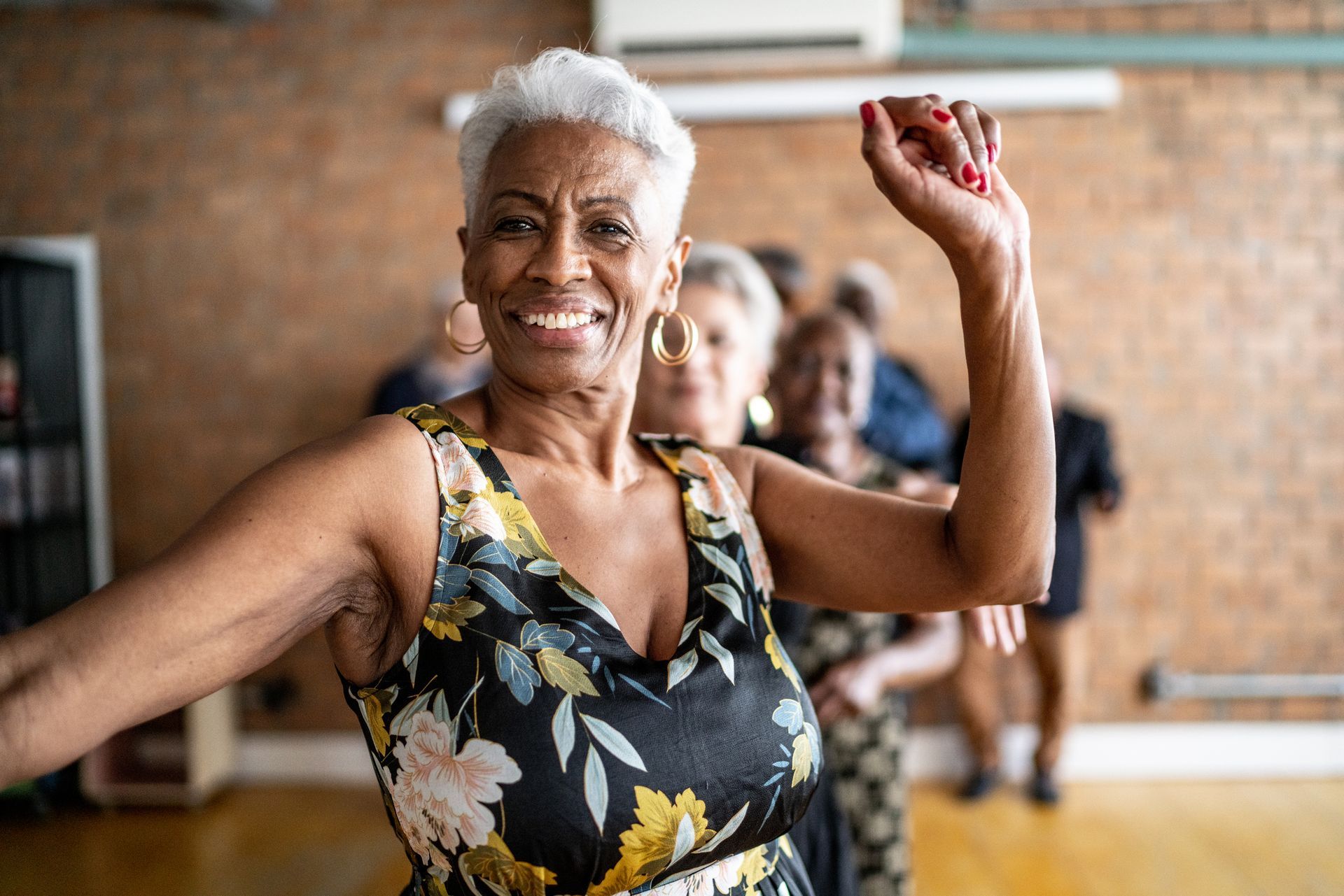 Smiling Black woman in floral dress dances in a group, arm raised, others behind her.