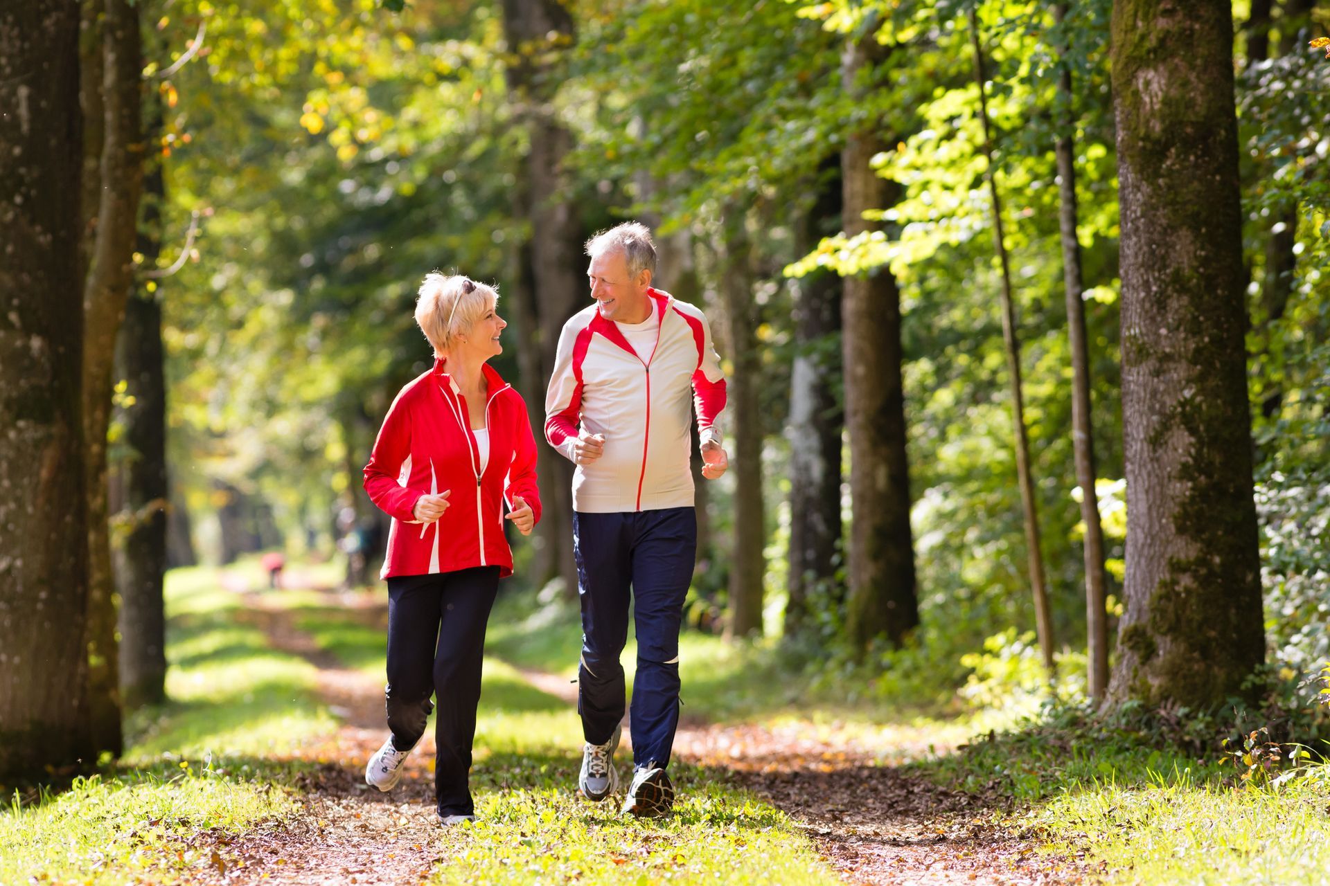 Elderly couple jogging on a sunny forest path, smiling.