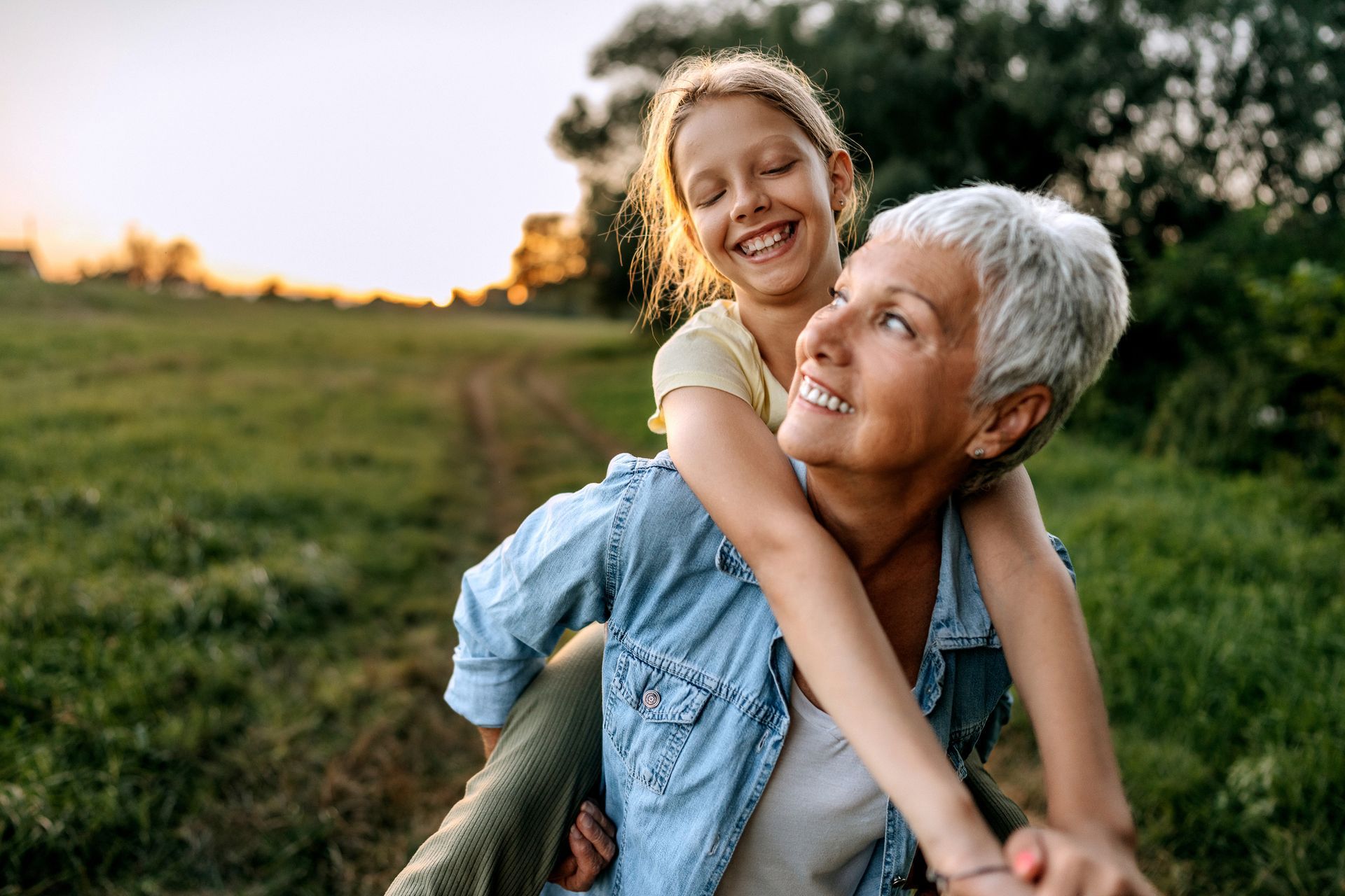 Grandmother giving a piggyback ride to a smiling granddaughter in a grassy field at sunset.