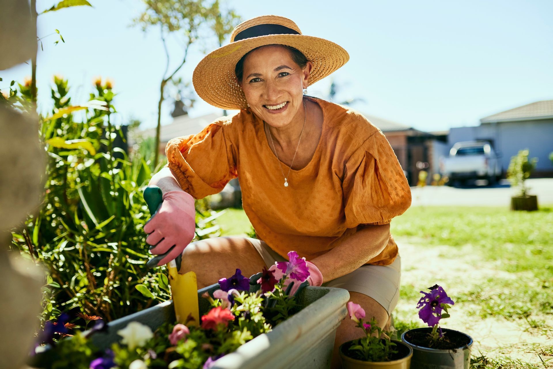Woman gardening, wearing a straw hat, orange shirt, and gloves, smiling in a sunny garden.