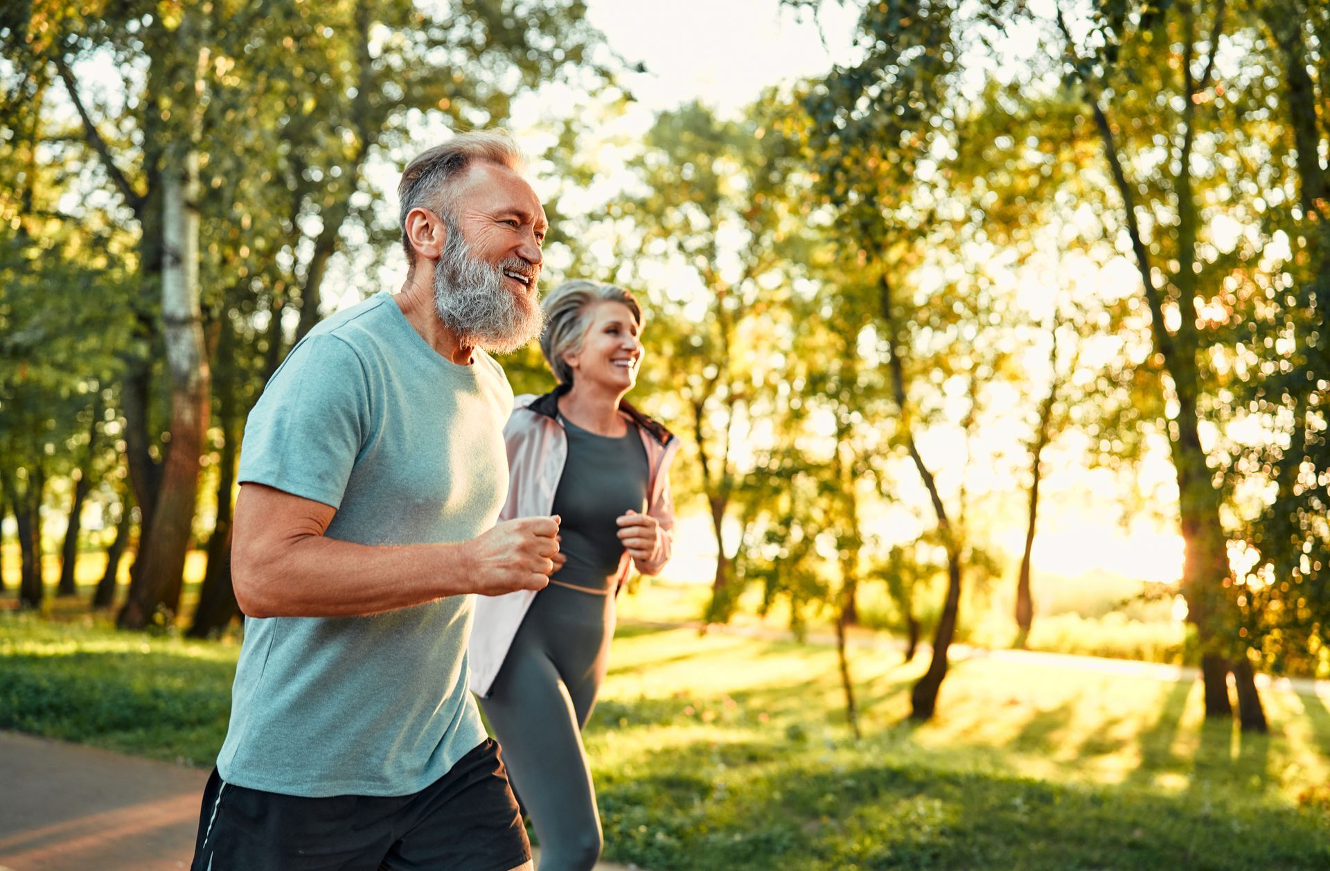 An older couple running happily on a path in a sunny park, surrounded by green trees.