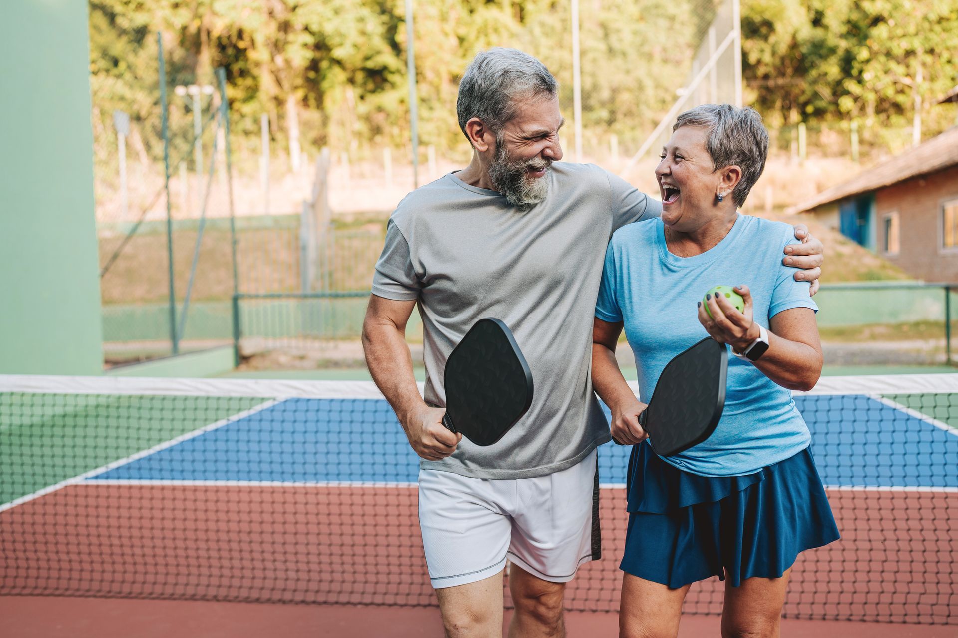 Happy senior couple, arms around each other, holding paddles and ball on a pickleball court.