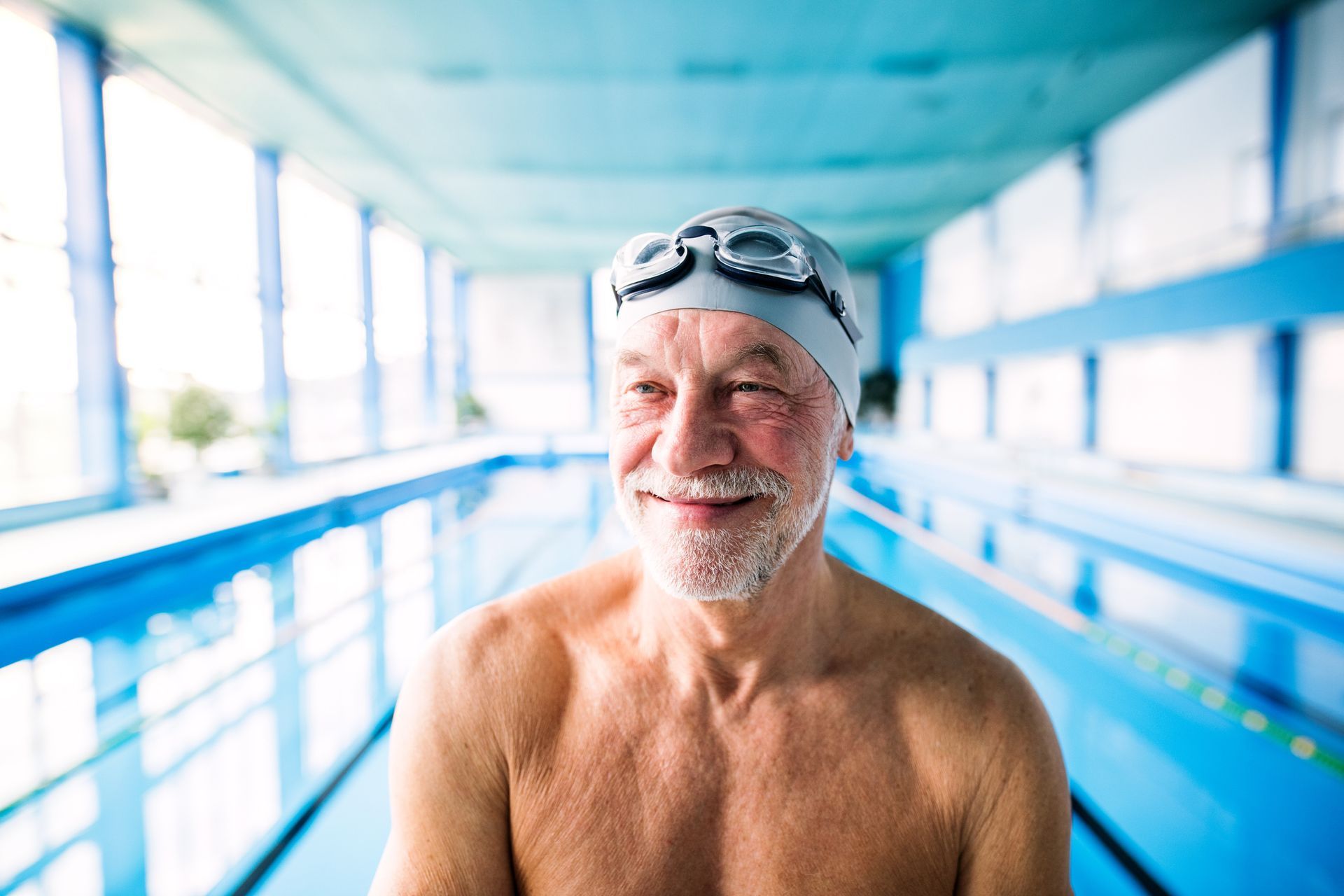 Smiling senior man in a swim cap and goggles stands in front of a blue indoor pool.