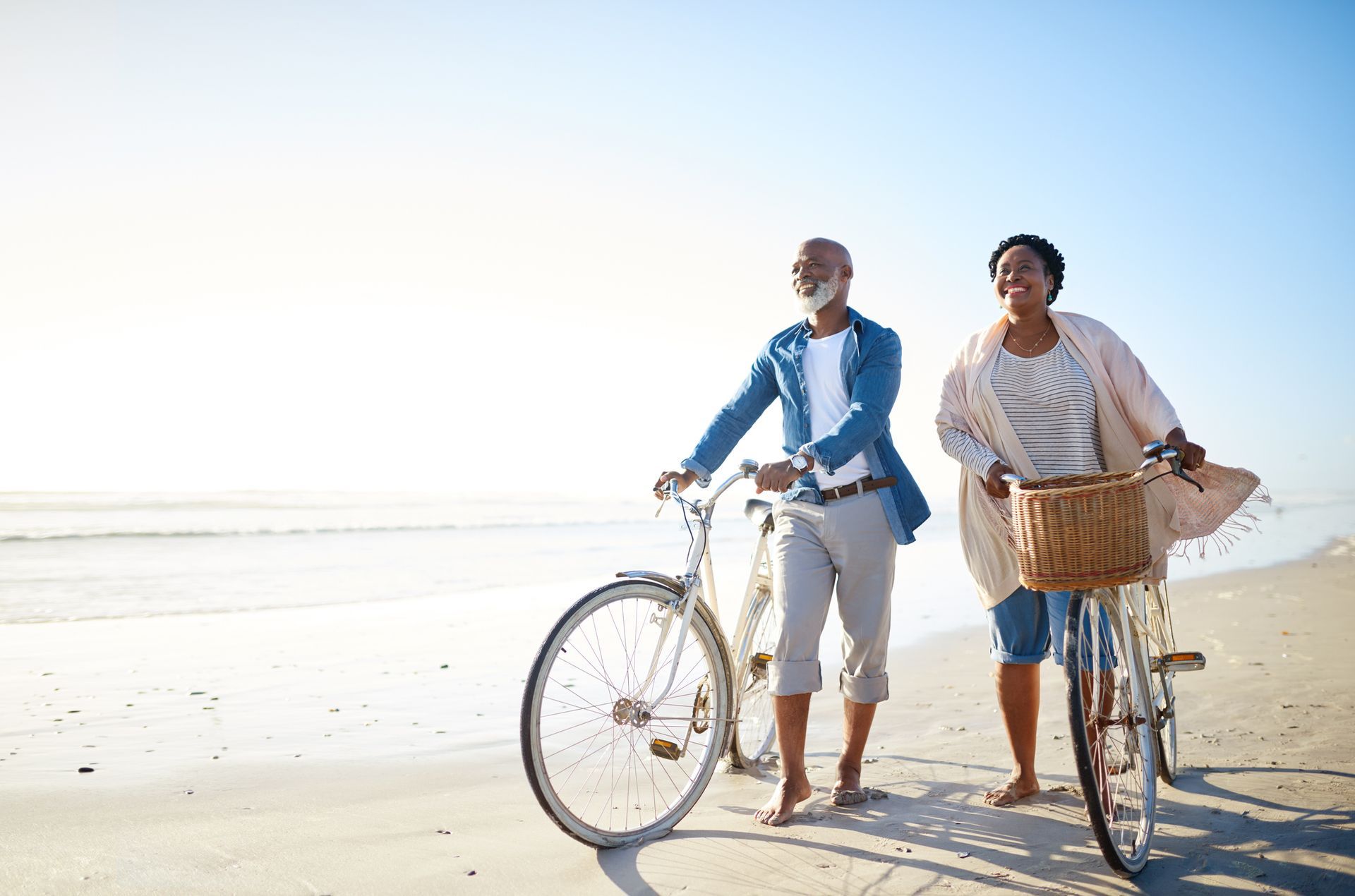Mature Black couple walking on beach, pushing bicycles.