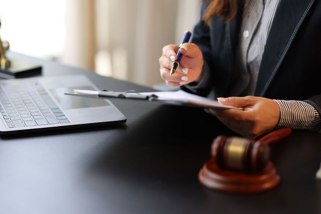 Hands writing on a clipboard beside a laptop and judge’s gavel on a desk Hands writing on a clipboard beside a laptop and judge’s gavel on a desk