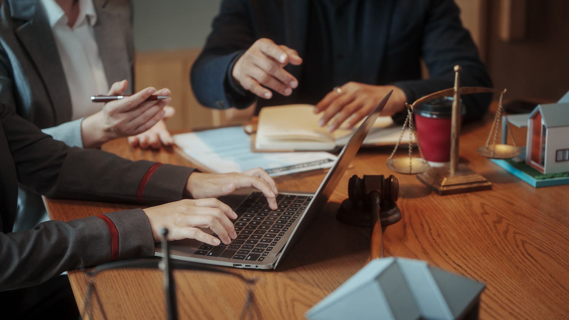 Colleagues at a wooden table discussing work around a laptop, notebooks, and coffee