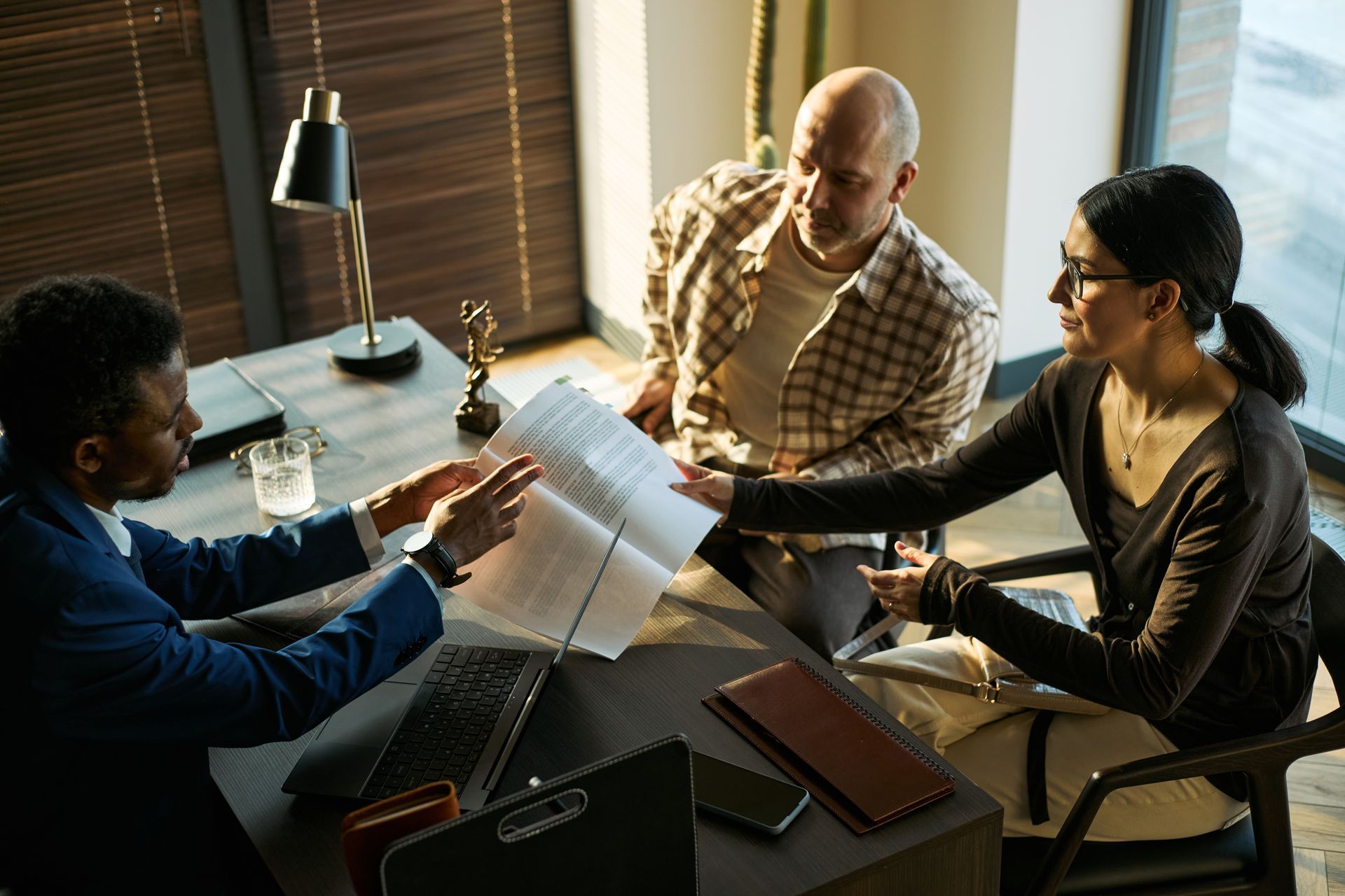 Three people reviewing documents around a laptop at a sunlit office table. Three people reviewing documents around a laptop at a sunlit office table.